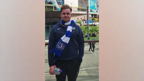 Lincolnshire Police Connor - a man with short brown hair which is going upwards - is wearing a navy jacket and a white and blue striped Chelsea scarf. He is standing in front of a stadium.