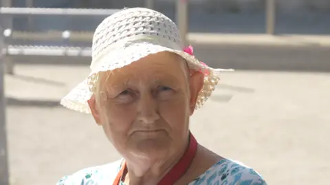 Jamie Niblock/BBC Linda Robinson is wearing a cream hat and a white and blue dress in front of a large concrete area.