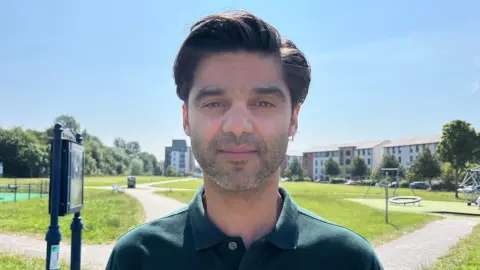 A man with dark hair, wearing a dark shirt standing in front of a children's playpark