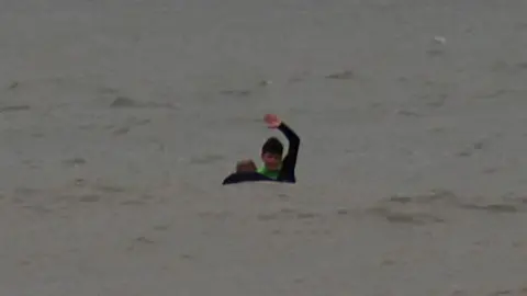 Neil Phillips Oscar is in a grey sea signalling with his arm over his head and the head and shoulders of a man is visible in the water next to him.