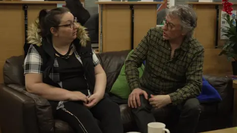 Steve Coogan sat on a brown leather sofa talking to a woman with long dark hair tied up in a ponytail, who is wearing clack trousers, a clack top, a white and black checked shirt and glasses.