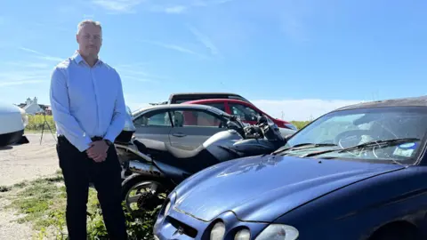 Deputy Andy Cameron in a blue shirt and smart black trousers stood in a coastal car park next to a car that has been abandoned and filled with rubbish