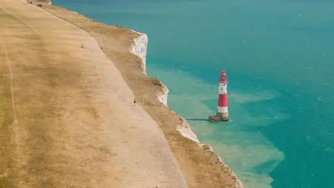 People walking on bone dry grass, along the cliff-top above the lighthouse at Beachy Head, near Eastbourne.