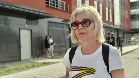 Blonde lady in her sixties wearing sunglasses stands in the street. The background is blurred but there are uniformed officers behind her