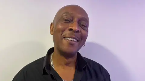 A man wearing a black shirt and silver chain smiles at the camera. 