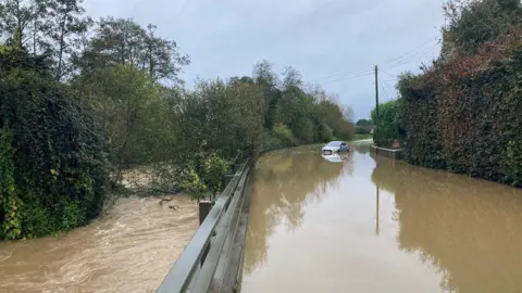 An A-road is flooded and a car is left stranded further down the carriageway. The river is overflowing with brown water surrounded by trees and bushes.