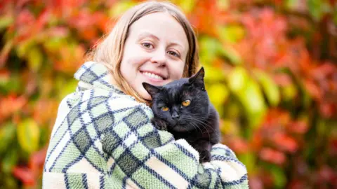 Joanne Booker has medium length blonde hair and is holding Panzer - who is black with orange eyes.
