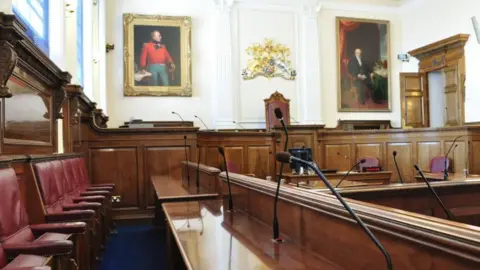 A view inside the states chamber. It shows red seats and dark wood stands. Large portraits of former states members are on the wall. 