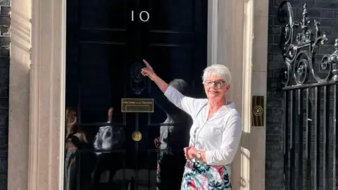 A woman with short white hair, glasses, a white shirt and floral skirt, stands and smiles outside the front door of Number 10 Downing Street while pointing up at the number on the door. 