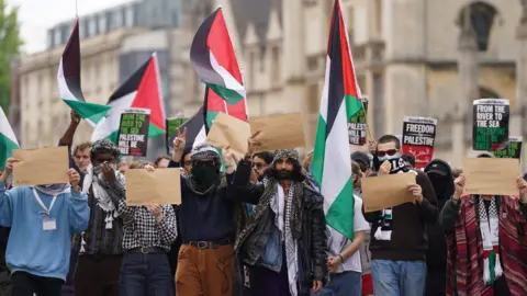 Students march to deliver their demands to Cambridge University as they protest against the war in Gaza. They wave Palestinian flags and hold up brown pieces of paper.