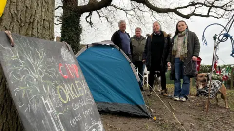 A chalkboard which says Camp Oakley to the left, a blue two man tent is in the middle of the picture, and four residents from Save Cowfold Oak standing on the muddy floor looking at the camera