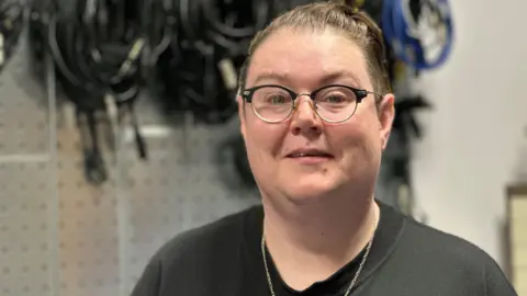 Sarah Callow, who has short dark hair and is wearing glasses and a black T-shirt. She has a silver chain around her neck and is standing in front of a rack of equipment leads.