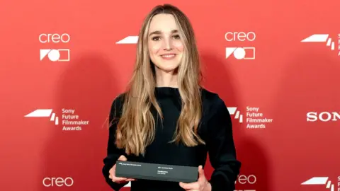 A woman in a black dress with long golden hair stands in front of a red background holding a black oblong box.
