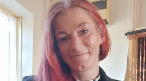 A slim woman with pink hair who is smiling at the camera. She is standing next to a window with bright light shining through. Her hair is tucked behind one ear and is over her other shoulder. 