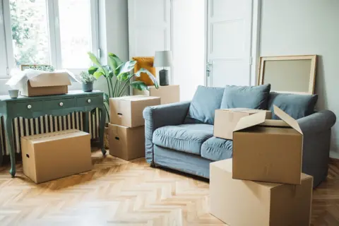 Getty Images Moving day concept, cardboard carton boxes stack with household belongings in modern house living room, packed containers on floor in new home.