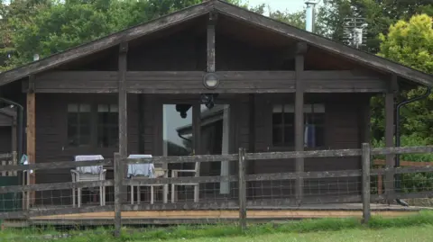 A log cabin holiday home, with chairs on a veranda outside it. There is grass in front and trees behind the cabin.