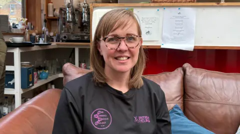 Laura has shoulder length light brown hair and round glasses and wears a black t-shirt with pink Cornwall Bicycle Project and British Cycling logos on it. She is sitting on a brown leather sofa in the cafe. 