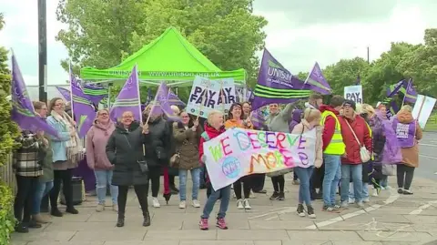 Picket line of striking healthcare workers with Unison banners and a colourful sign