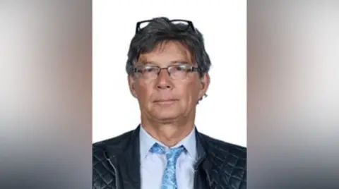 Barry Martin wears a blue tie and shirt and two pairs of glasses, one perched on top of his head. 