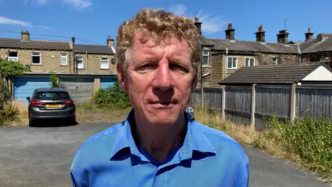 A man with blond, curly hair and wearing a blue shirt standing outside with a row of terraced houses behind 