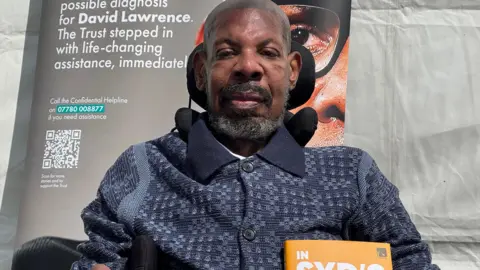 Syd sitting in a wheelchair with a dark polo shirt on, short beard and hair, smiling at the camera. There is a poster behind him and he is outside by a white tent.