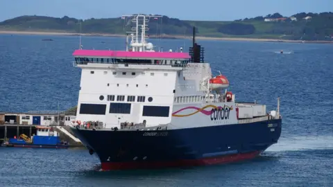 A white and blue Condor Ferries boat enters the harbour in St Peter Port, Guernsey. It is a sunny day and the sea looks calm. The boat has just gone past the harbour wall.