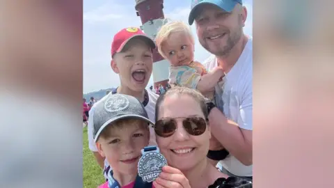 Jennifer Hooper Steve and Jennifer Hooper and their three children are standing in front of Smeaton's Tower lighthouse on Plymouth Hoe. They are showing off a medal won by one of the children. All are smiling at the camera in the sunshine