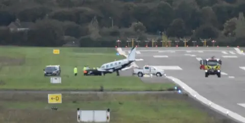 Pete Smith A picture of the runway at Gloucestershire Airport. The plane can been seen on the grass, with the nose slightly pointed down. At least two vehicles appear to be next to the plane. 