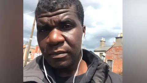 A man looks into the camera with a neutral expression. His name is Yves Sakila, originally from the DR Congo. He is outside on a street, in the background terraced houses can be seen along with a cloudy sky. He is wearing a black coat and has white wired earphones in his ears. 