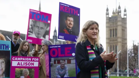 A woman with blonde hair - Kim Leadbeater - stands at Parliament Square in London in front of a crowd of pro-assisted dying campaigners holding purple and pink placards with pictures of different people's faces on.