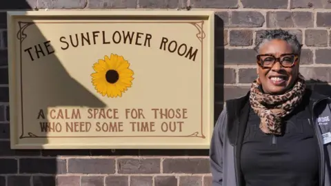 Community engagement officer Andrea Nelson stands by a sign for the new space, the Sunflower Room. The sign has a picture of a sunflower and a sign that says "a calm space for those who need some time out".