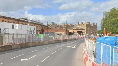A road with barriers and construction work on both sides. A Victorian railway station building on one side of the road in the distance. A tree on the other side.