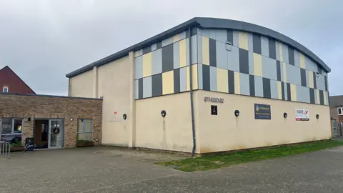Carmelo Garcia A photo of the outside of a community centre with cream walls and yellow, blue and grey cladding