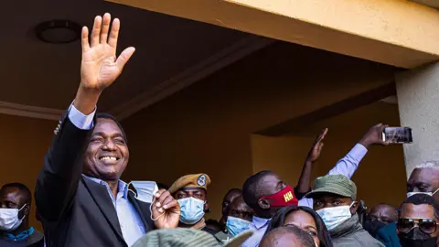 AFP via Getty Images Hakainde Hichilema in an open blue shirt and dark blazer smiles and waves at his supporters.