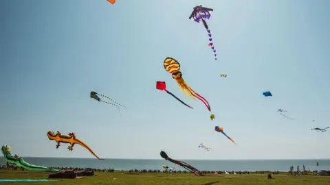 Visit Hull and East Yorkshire A number of colourful kites fly over a large grassed area on a clifftop. The North Sea is visible in the background and is glistening under sunshine and a blue sky.