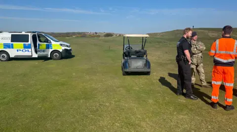 A police officer, an army officer and another man wearing orange high-vis clothing stand on a green golf course. They are in conversation. There is a golf buggy and a police van to the back of them. It is a sunny day. 
