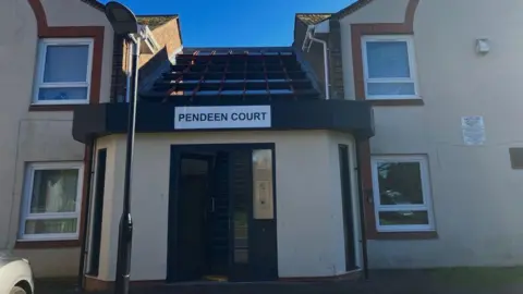 Picture of Pendeen Court it has a sign on the top of the door saying Pendeen Court. There is a glass door which is open ajar by a cleaners sign. 