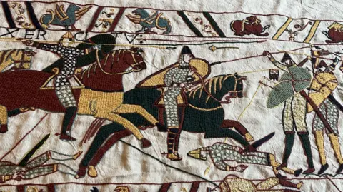 John Devine/BBC A piece of tapestry which shows horses and men with shields and weapons.