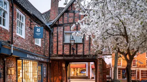 Getty Images A red brick, half-timber building in York, with a National Trust gift shop below. In the right corner, a tree with white cherry blossom can be seen. 