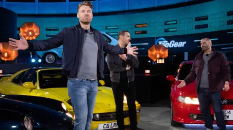 Flintoff with arts outstretched on the top Top Gear set, in front of Paddy McGuinness and Chris Harris and two cars