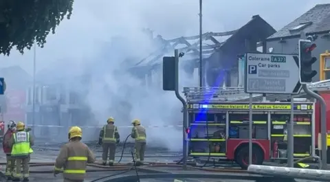 Fire-fighters behind a cordon tackling a fire in a derelict building, with a fire engine to their right and smoke emanating from the building in front of them 