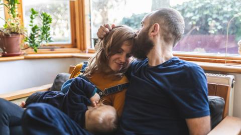  stock photo of a woman on the sofa breastfeeding her little son and sharing time with her husband
