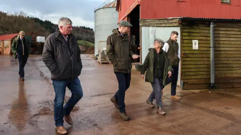 Kensington Palace The Prince of Wales, wearing blue jeans, walking boots, a green waterproof jacket and a grey flat cap, is walking next to a barn. He is talking to a woman who is walking with him as three other men walk alongside.