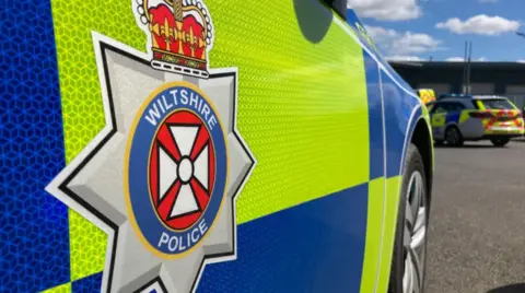 The side of a police car with a Wiltshire Police logo and blue and yellow coloured panels