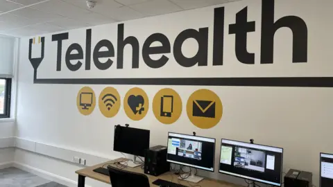 University of Suffolk A black printed sign on a wall says "Telehealth" and there are computers and monitors on a desk in the room.