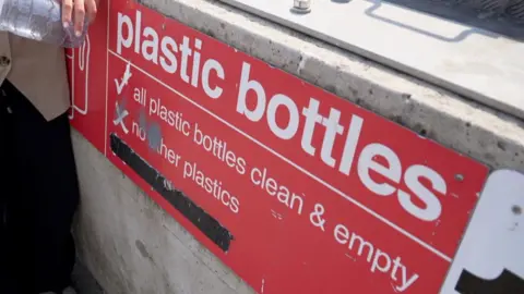 A red sign saying 'plastic bottles' in white writing. It is attached to the side of a bring bank at the La Collette recycling centre in St Helier. 