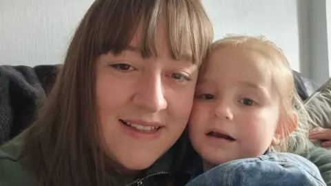 Murray family Dorothy Murray sitting on a sofa holding her young daughter Sienna.  Ms Murray has long, straight brown hair with a fringe and is wearing an olive green zipped jacket. Sienna has fair hair tied back. They are both looking into the lens. 
