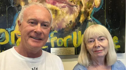 A middle aged man and woman are looking straight at the camera, you can see their head and shoulders and the bottom of a large painting is the backdrop. It has a plate on it saying "Chris Parry attended this school". The man has short grey hair and blue eyes and is wearing a white t-shirt. The woman has shoulder length blonde hair, greenish eyes and is wearing a blue tshirt and a silver necklace.