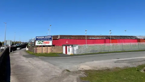A general view of Borough Park. The ground is next to a main road and is covered in sheets of corrugated metal, some painted red and some rusty. There are advertising boards to one side and a security fence to the other.