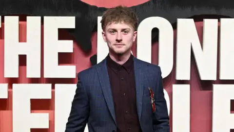 Locke wears a brown shirt sunder a blue tartan jacket, with sunglasses hanging form the suit jacket's pocket